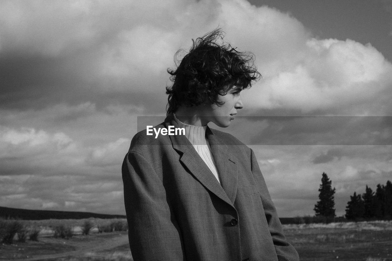 FULL LENGTH OF YOUNG WOMAN LOOKING AWAY ON FIELD AGAINST SKY