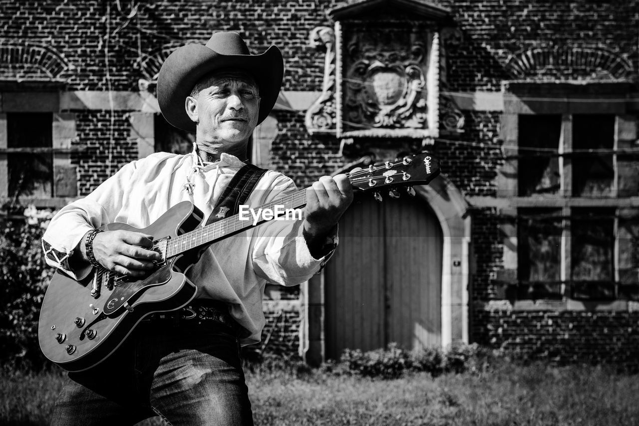 Man wearing hat playing guitar while standing against built structure