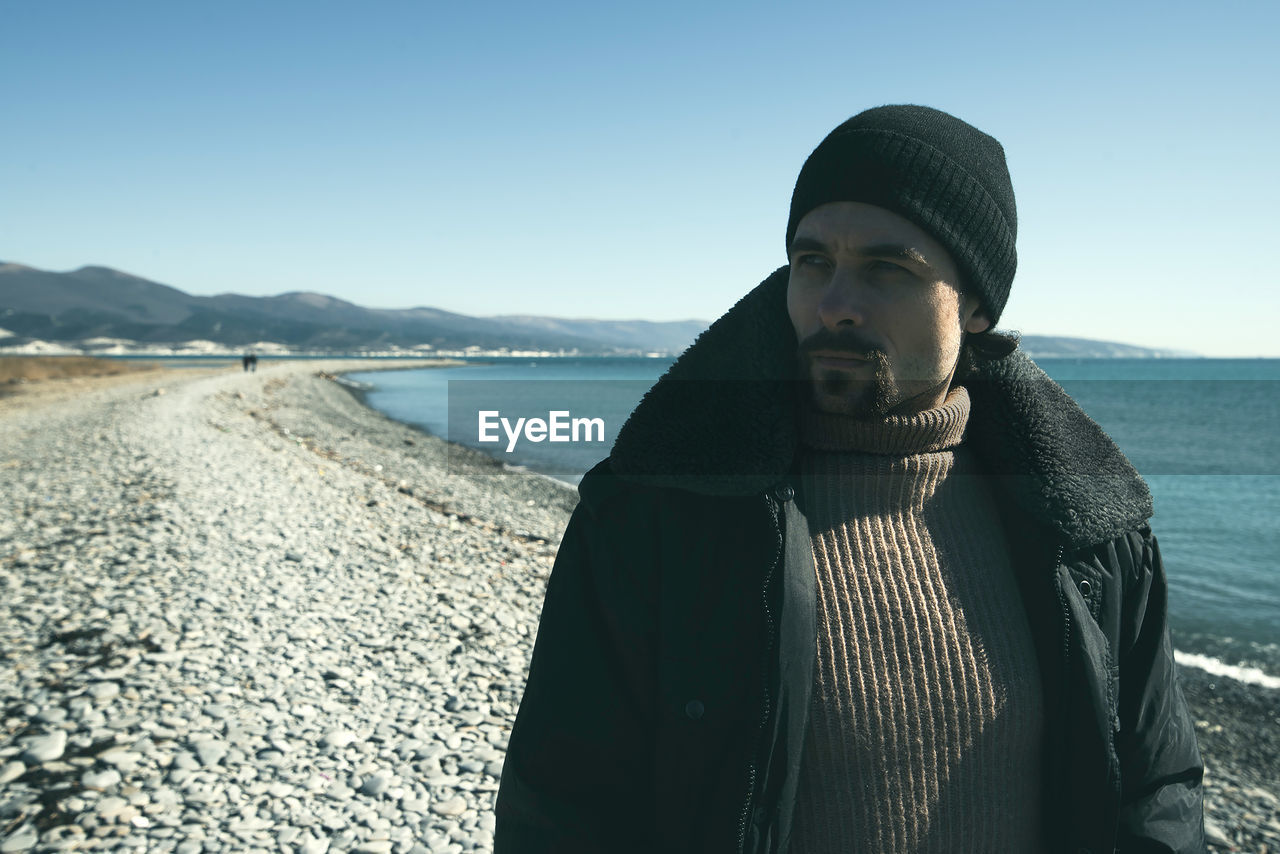 Thoughtful young man standing at beach against clear sky