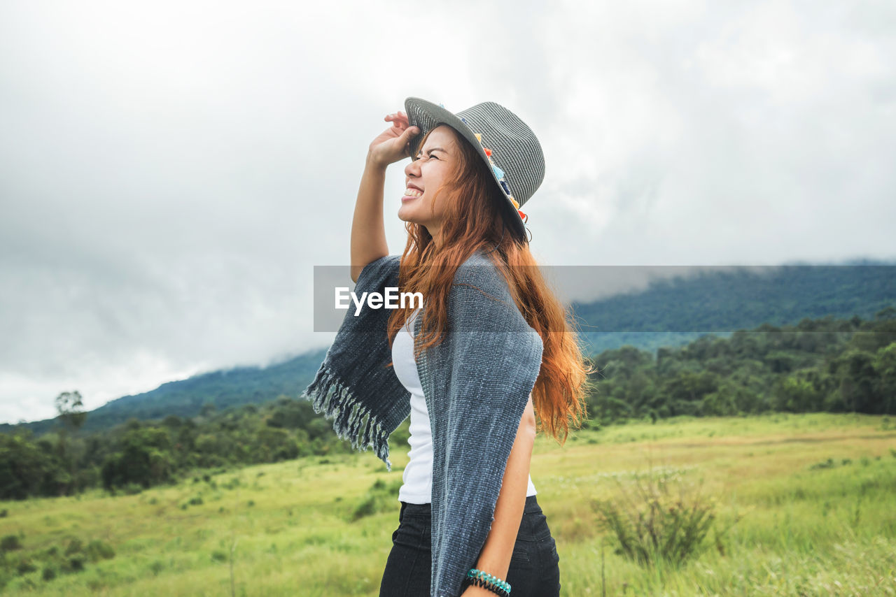 Side view of smiling woman wearing hat standing on field against sky