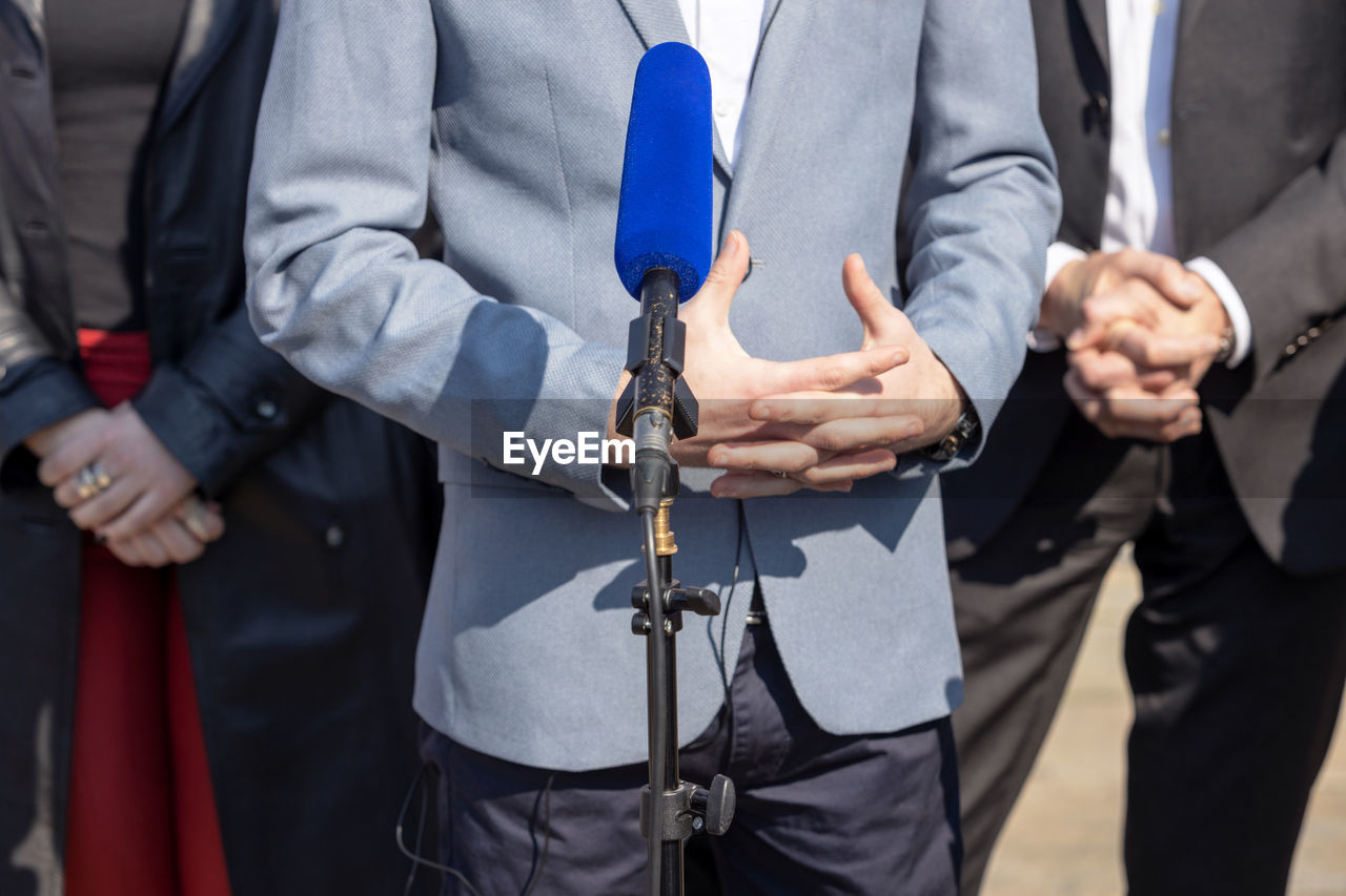 Microphone in the focus at news conference, midsection of man holding hands in the background