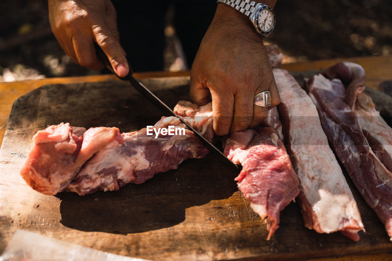 Crop unrecognizable man with watches and ring on finger butchering meat with knife on wooden cutting board preparing food outdoors