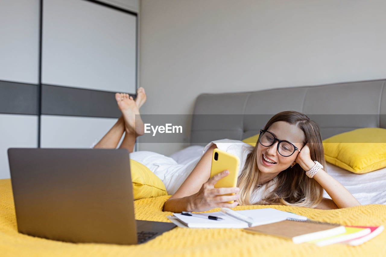 Smiling woman using phone while lying on bed at home