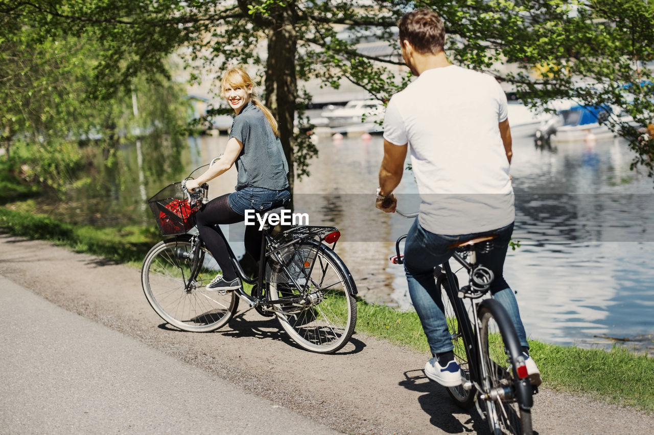 Happy businesswoman looking at colleague while riding bicycles on street by lake
