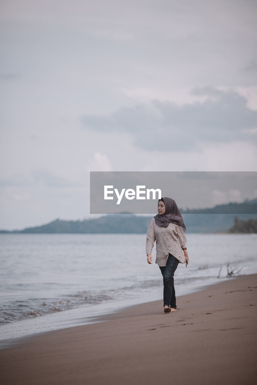 Full length of woman in hijab walking at beach against sky