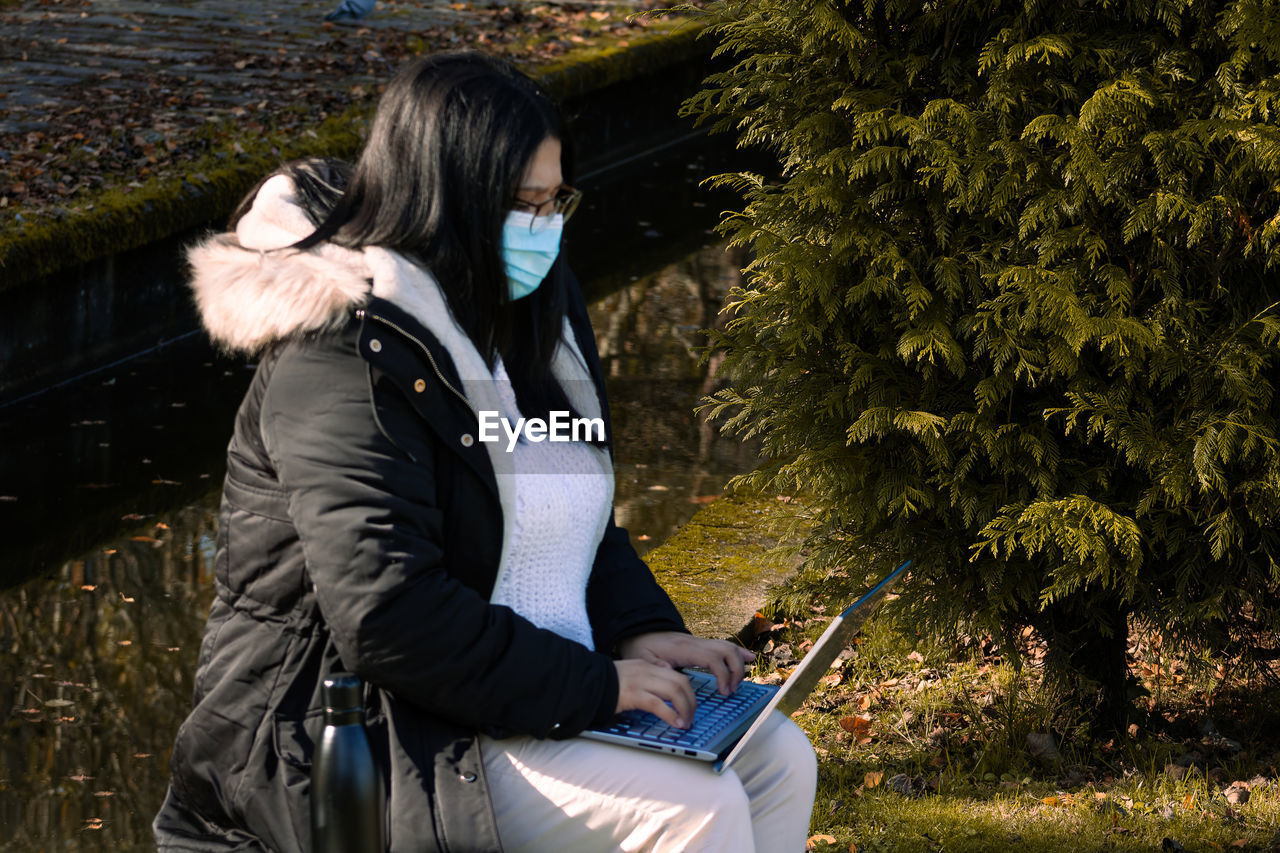 REAR VIEW OF WOMAN USING MOBILE PHONE WHILE SITTING ON PLANT AT PARK