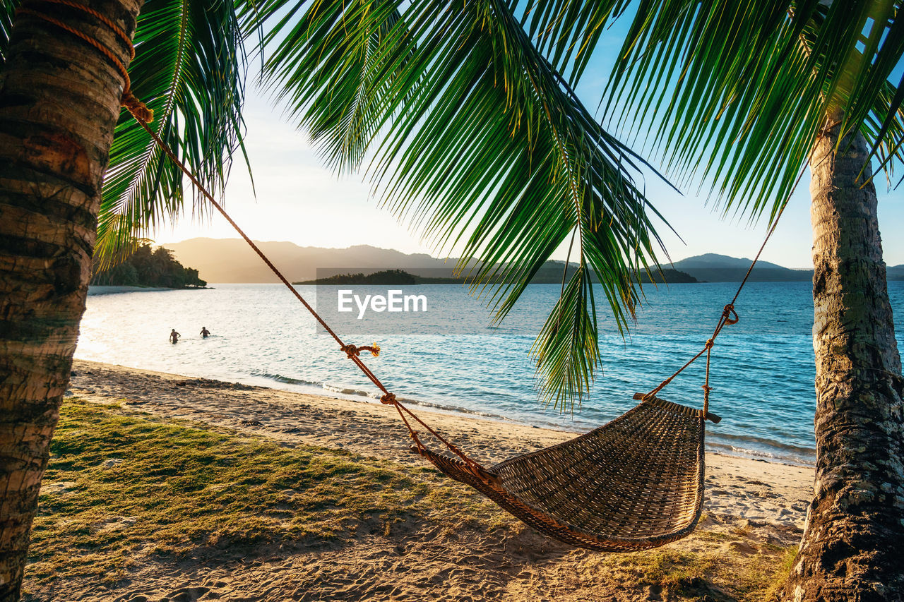 Empty hammock between two palm trees on beach at sunset. silhouette of couple in background in sea. 