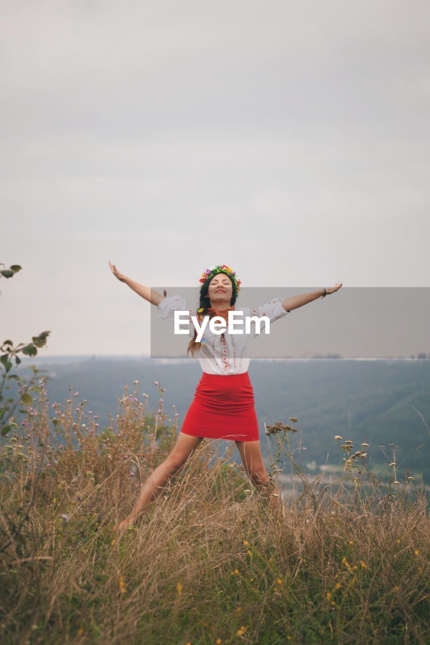 Smiling young woman with arms raised standing on field against clear sky