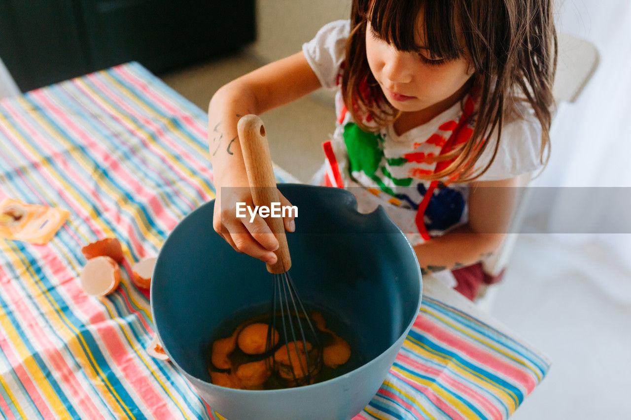 Little child whisking eggs in a blue bowl at the kitchen table