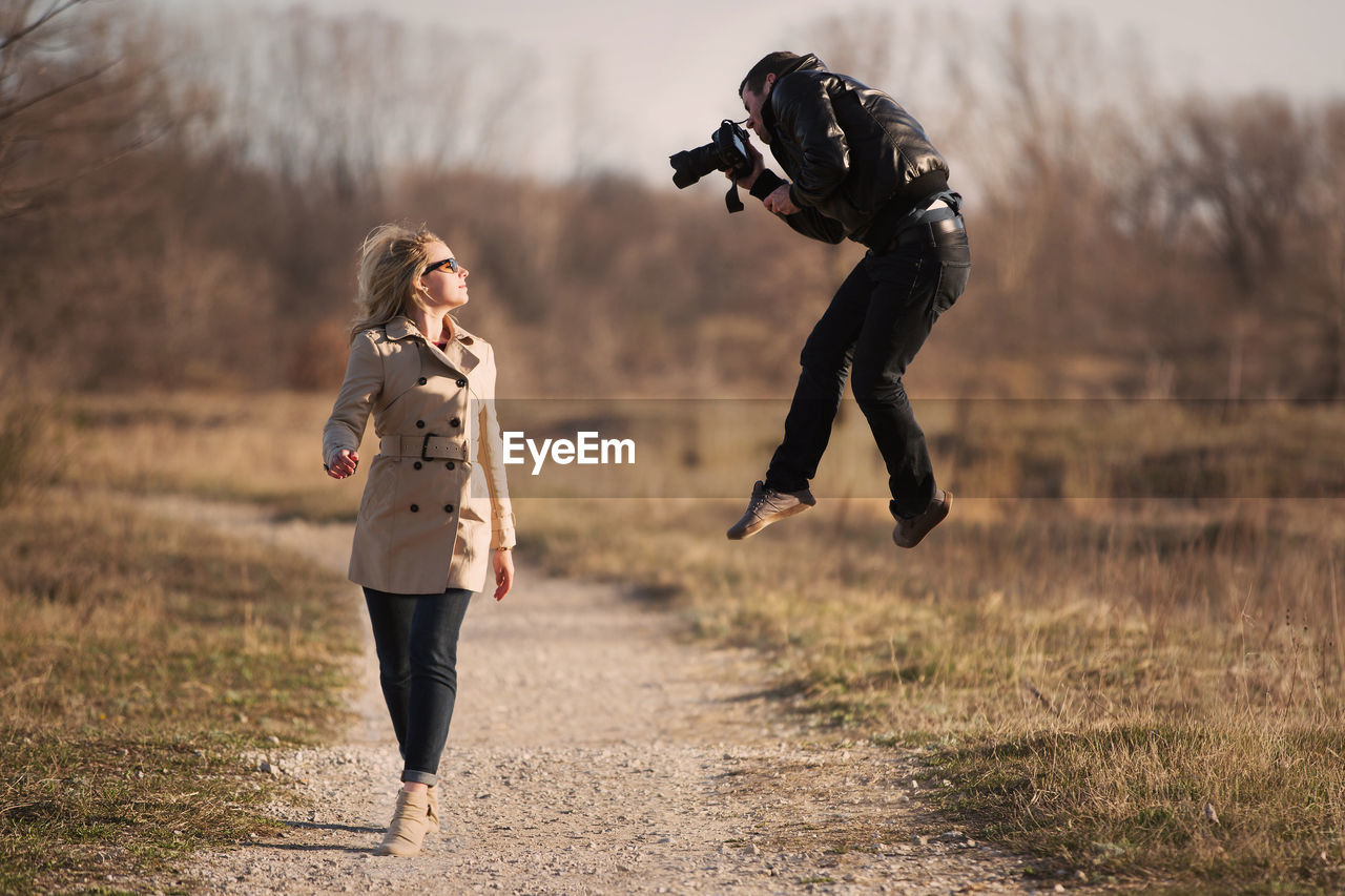 Full length of woman walking while photographer jumping on field