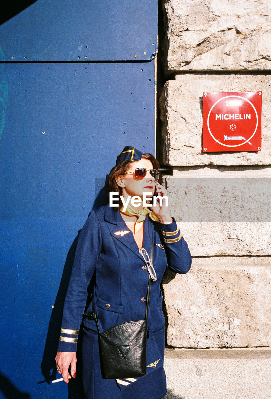 YOUNG WOMAN USING PHONE WHILE STANDING AGAINST WALL