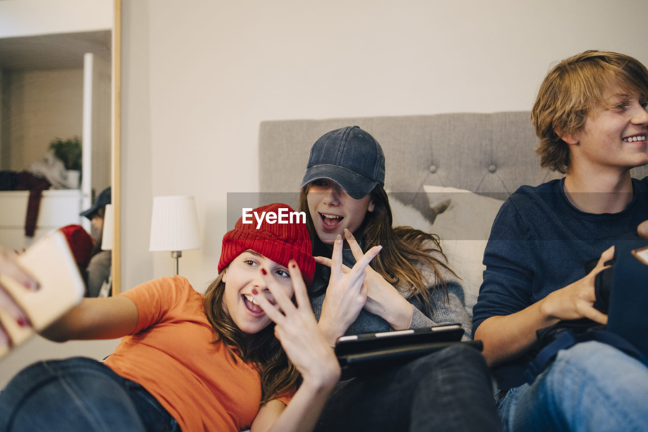 Girls taking selfie while sitting with male friend on bed at home