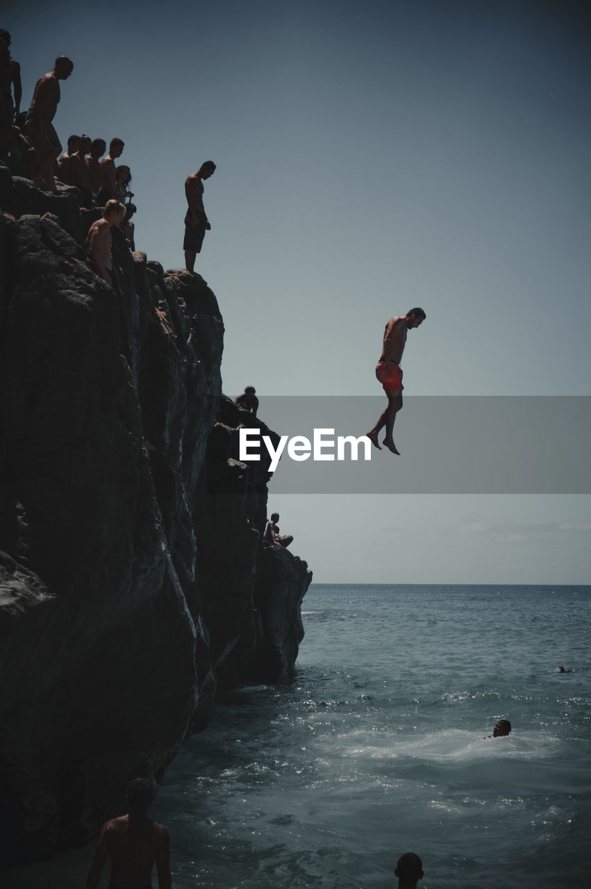 PEOPLE JUMPING ON ROCK FORMATION AT SEA AGAINST SKY