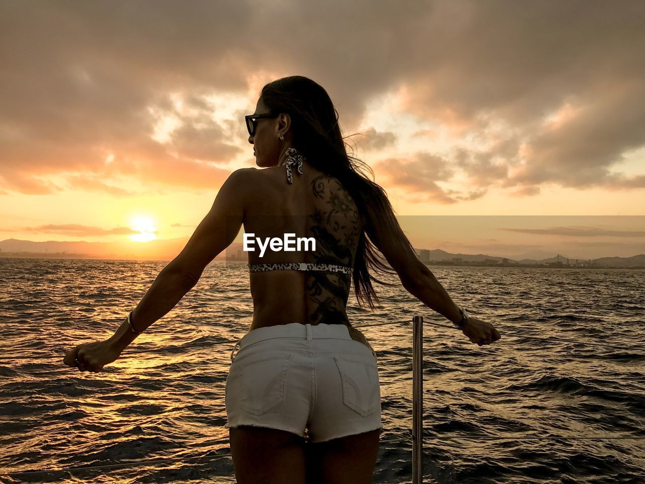 Rear view of young woman standing against sea during sunset