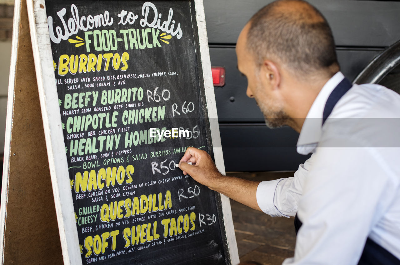 Close-up of male vendor writing on blackboard while crouching at street by food truck