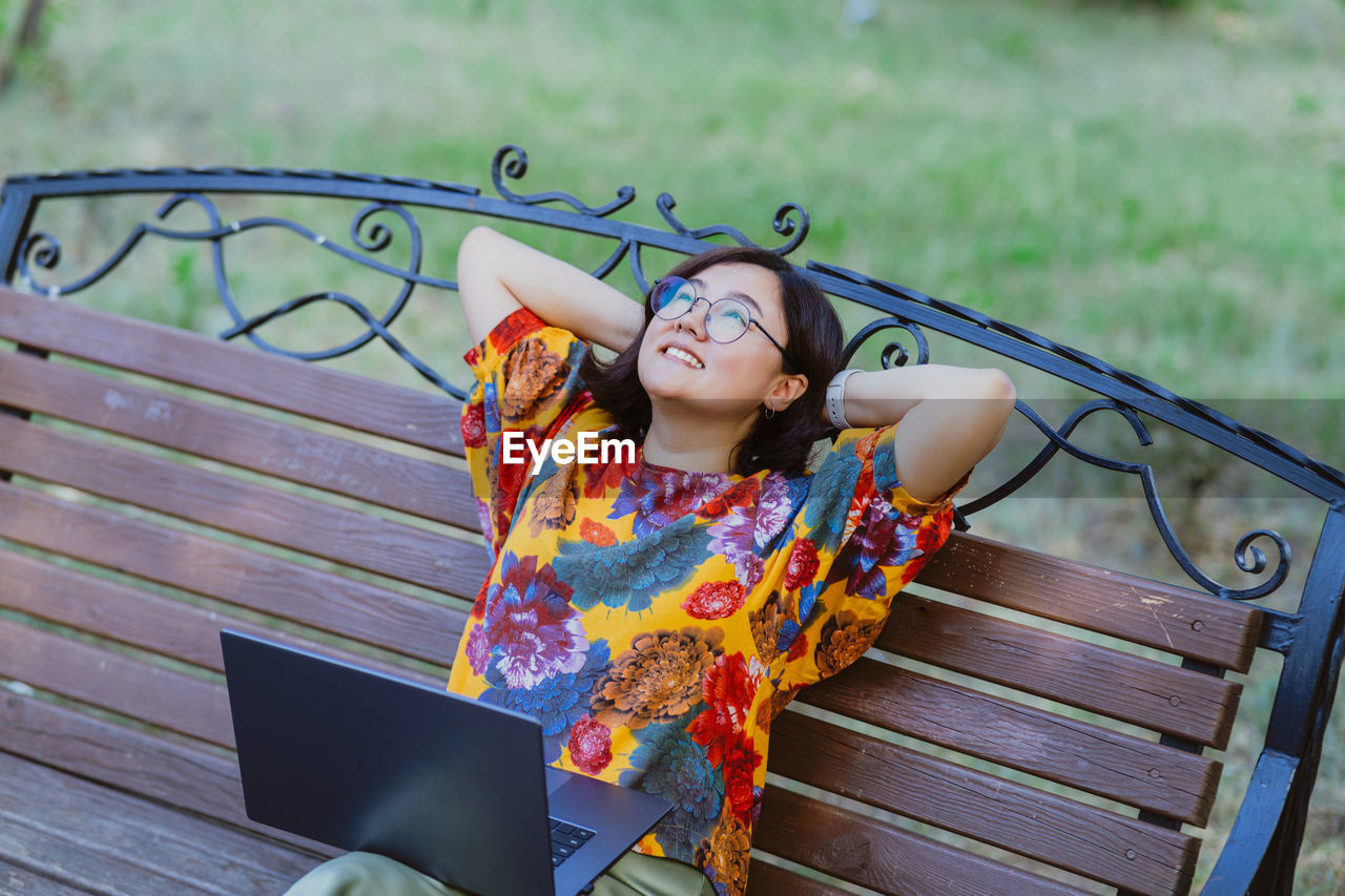 Asian freelancer with laptop enjoying relaxing on bench in park in summer