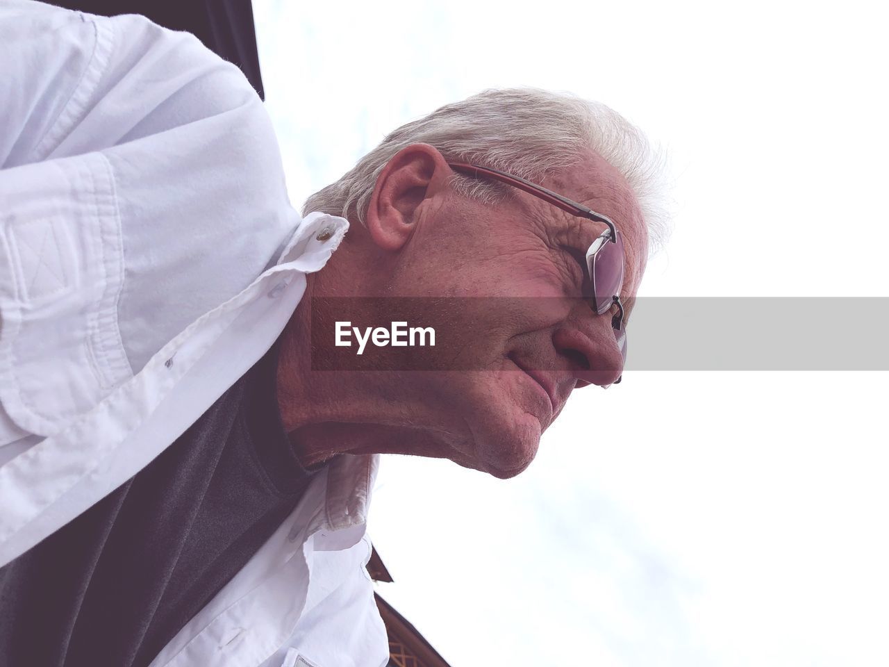 Low angle view of senior man wearing eyeglasses looking away while sitting against sky