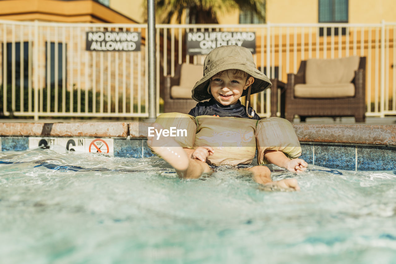 Front view of young toddler boy sitting in hotel pool on vacation
