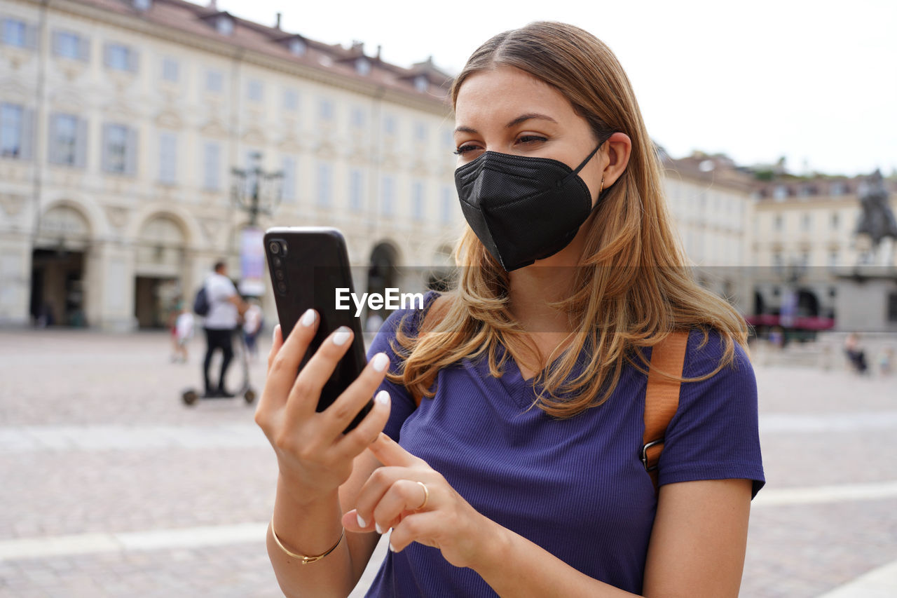 Portrait of young woman with black protective mask ffp2 kn95 using mobile phone with city background