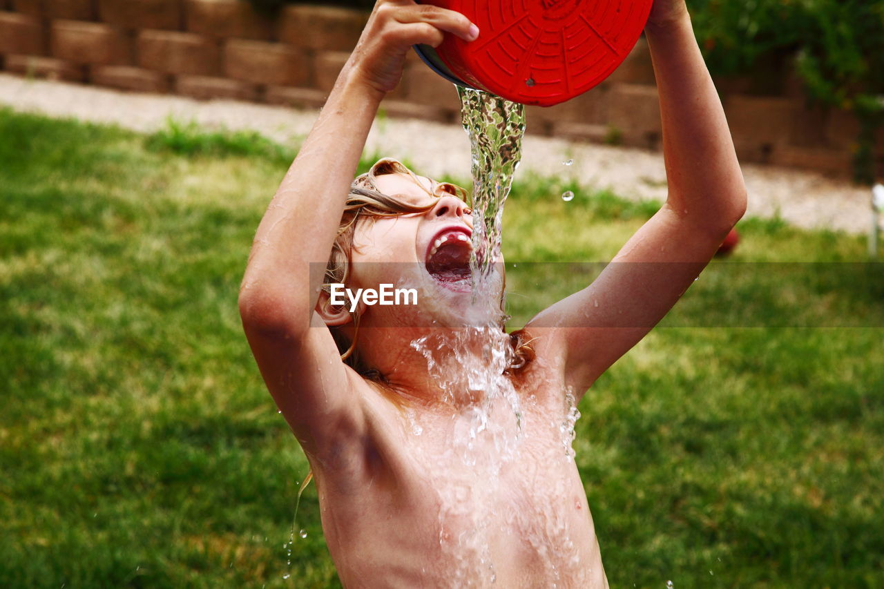Shirtless boy pouring water on mouth from container at lawn