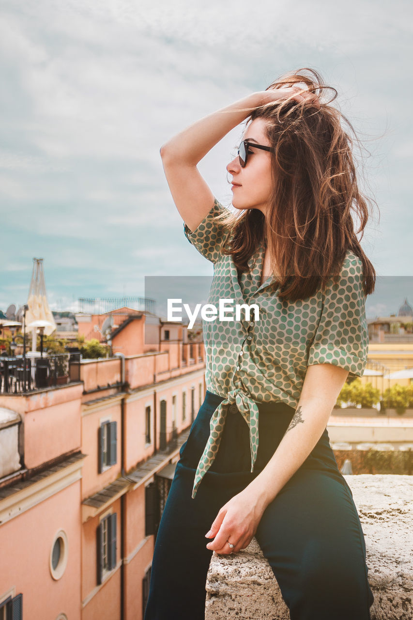 A young smiling woman in sunglasses sits on a rooftop with a panoramic view of rome, italy
