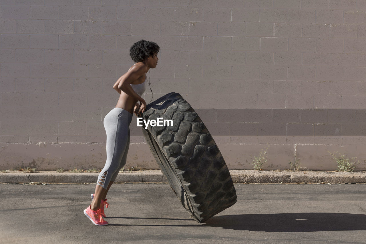 Full length of woman lifting tire truck while exercising against wall