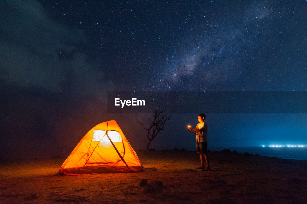 Side view of man standing by illuminated tent at beach during night