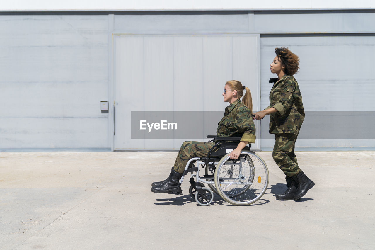 Young female army soldier pushing colleague on wheelchair at military base during sunny day