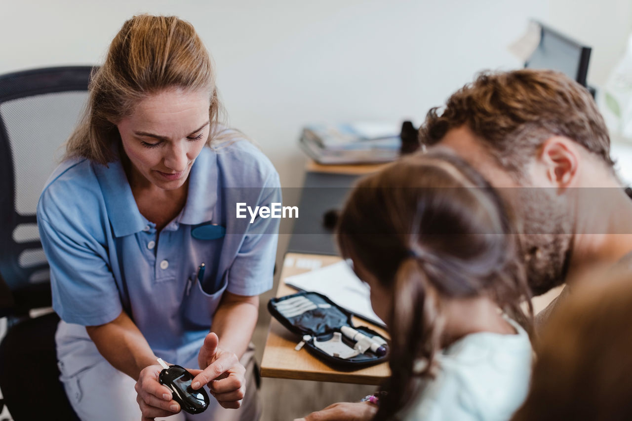 Female pediatrician showing glaucometer to girl sitting with father in hospital