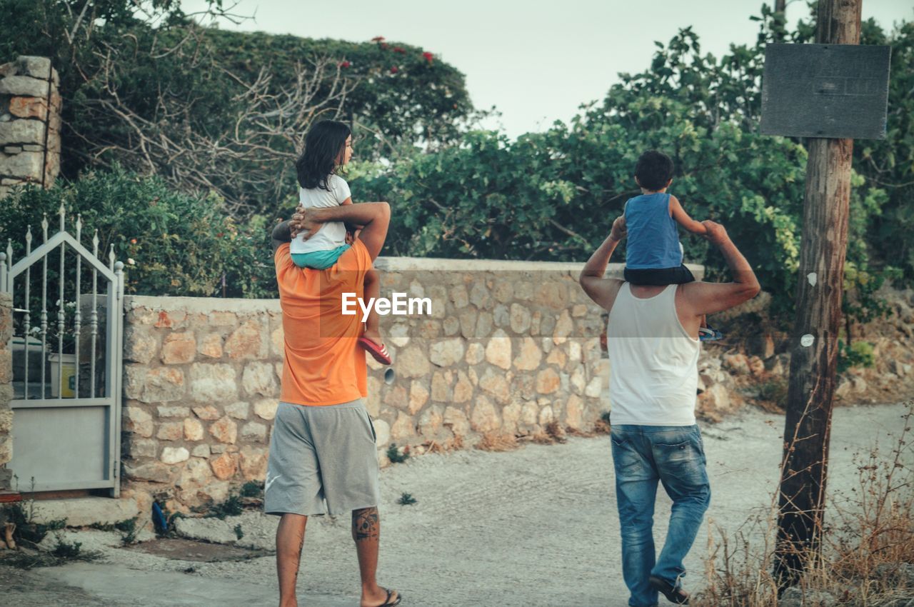 Rear view of men carrying children on shoulder while walking on walkway