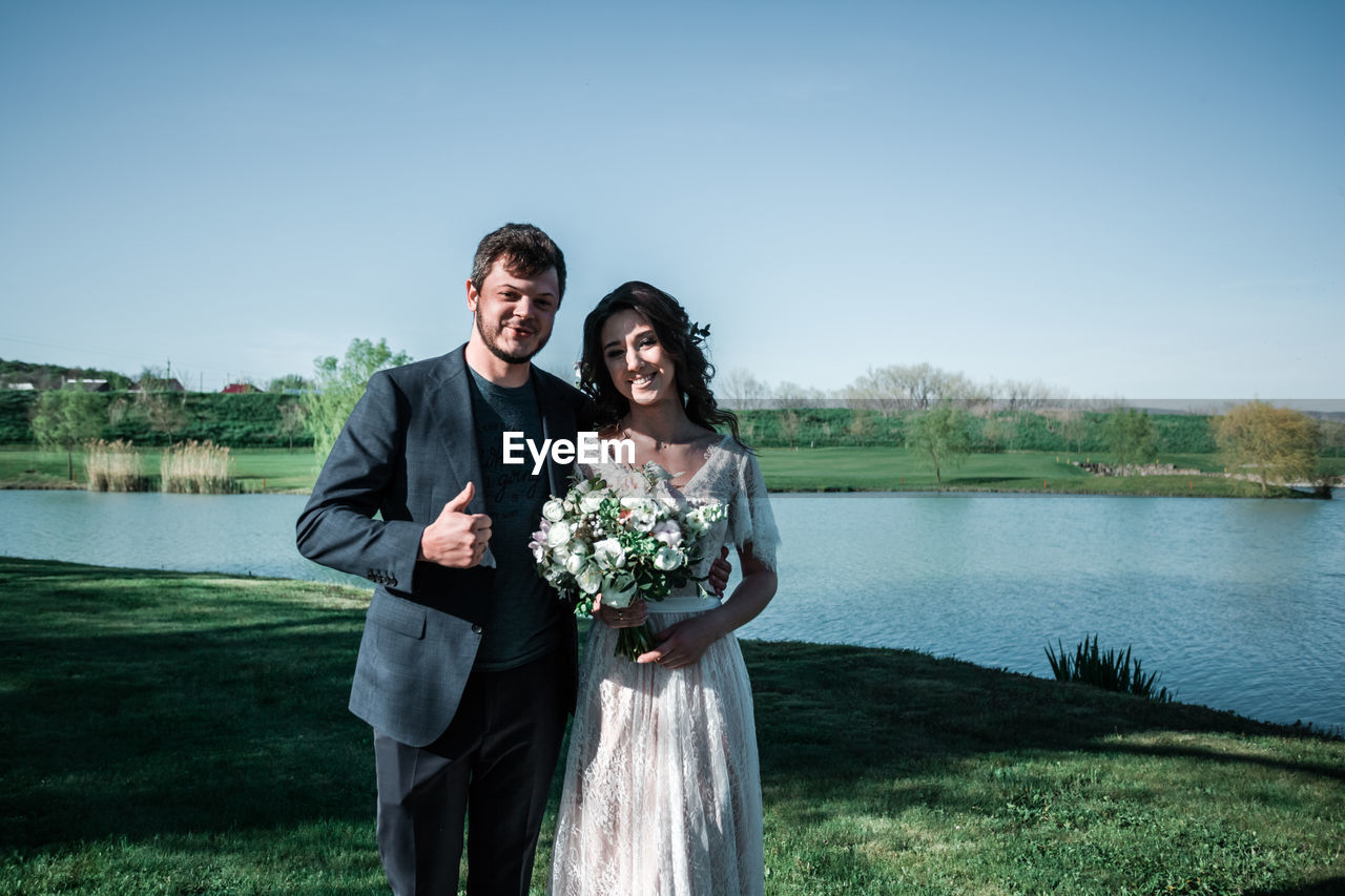 PORTRAIT OF SMILING YOUNG COUPLE STANDING ON LAKE