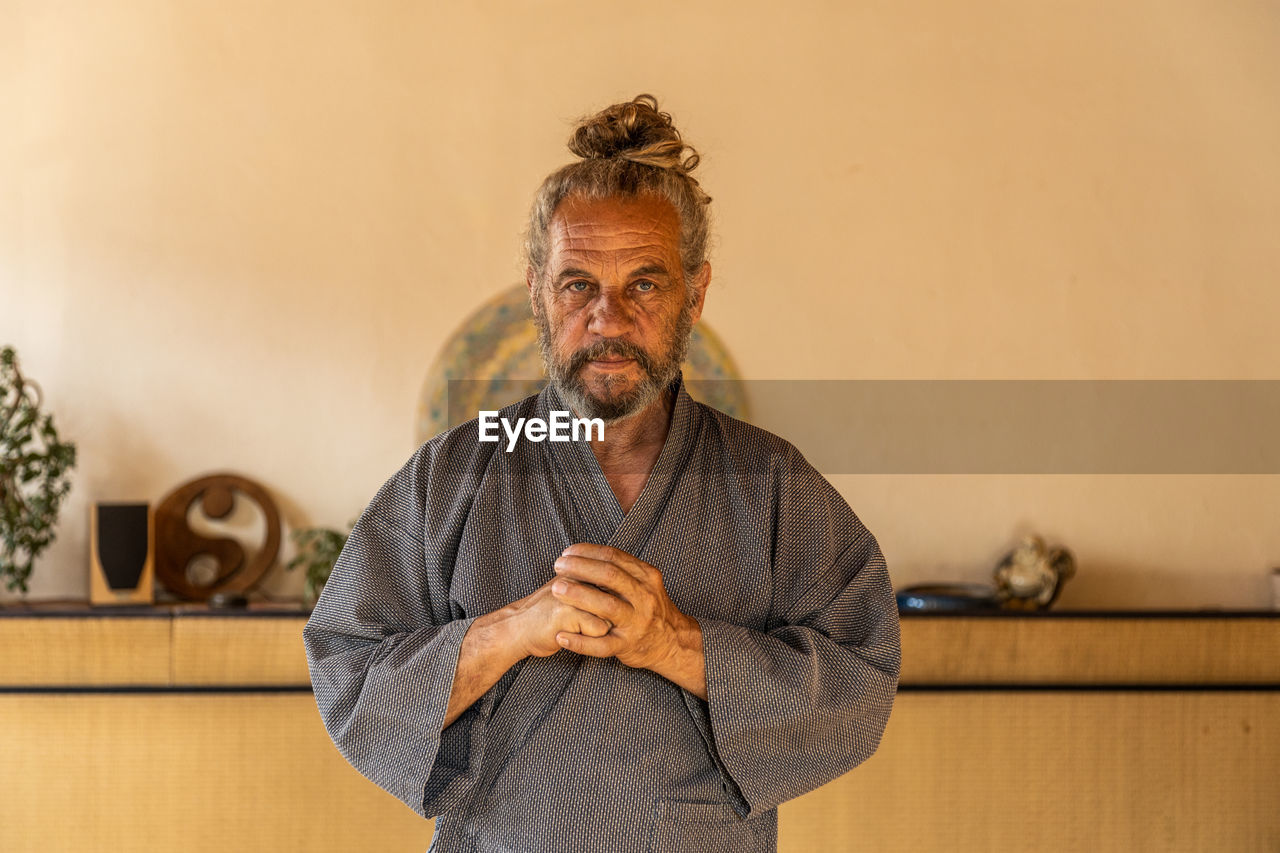 Senior bearded ethnic male chi kung practitioner in traditional wear looking at camera while standing in light studio