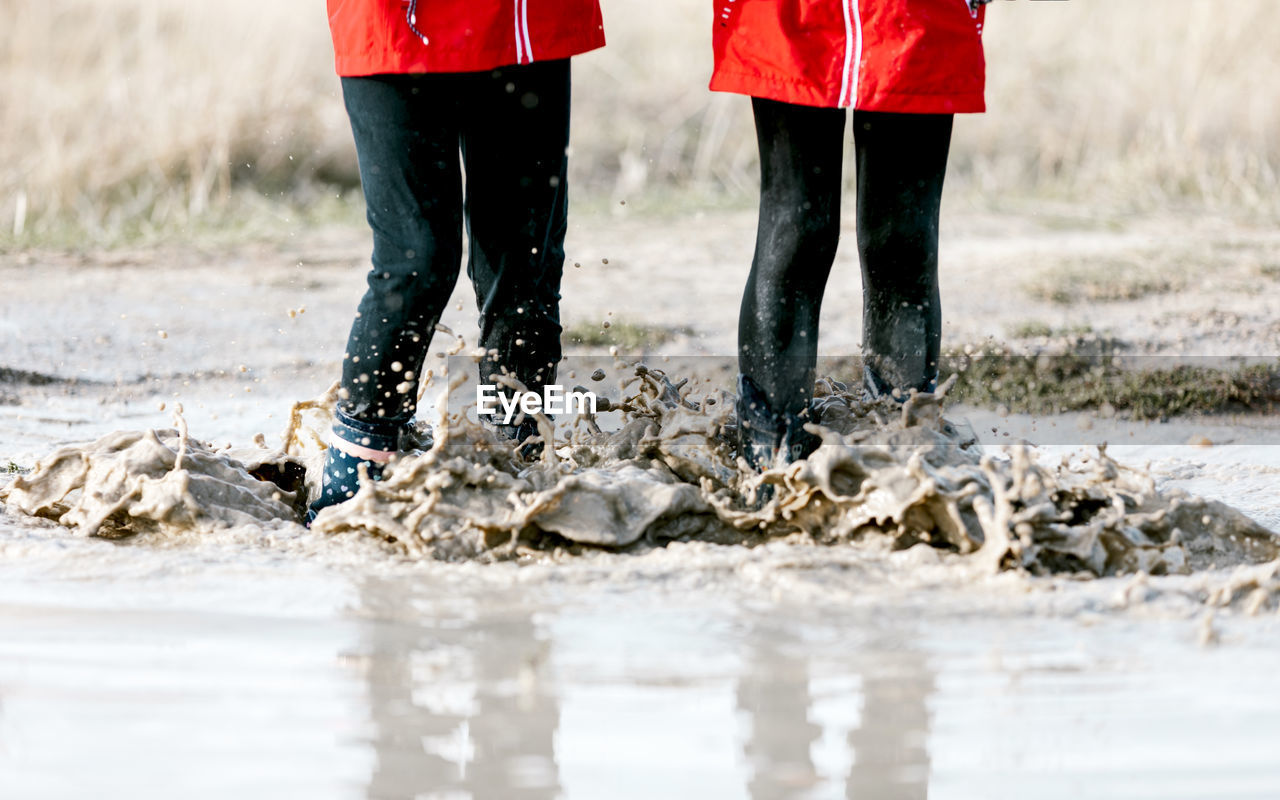 Unrecognizable carefree kids in raincoats and rubber boots splashing water in puddle while having fun on sunny day in countryside