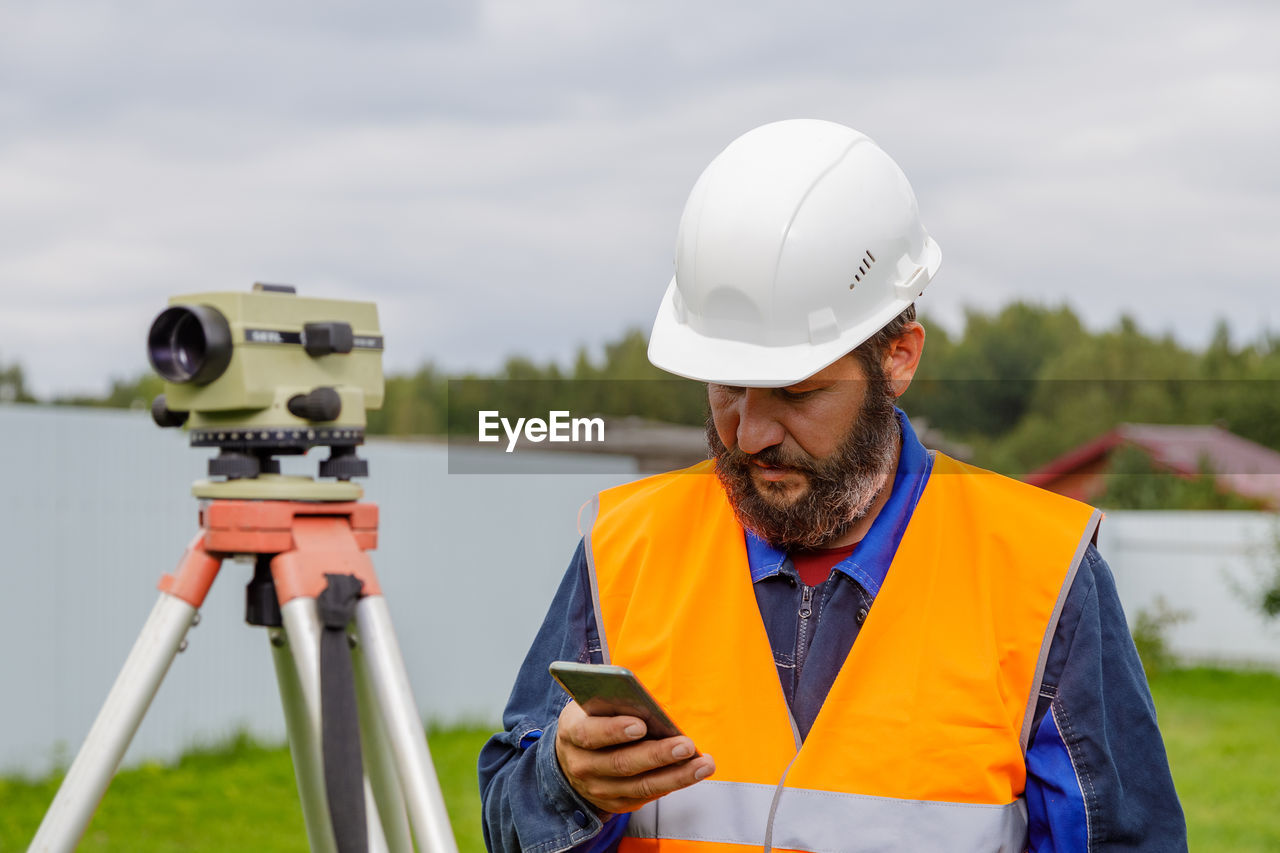 A civil engineer with an optical level looks into a mobile phone. a bearded man is looking 