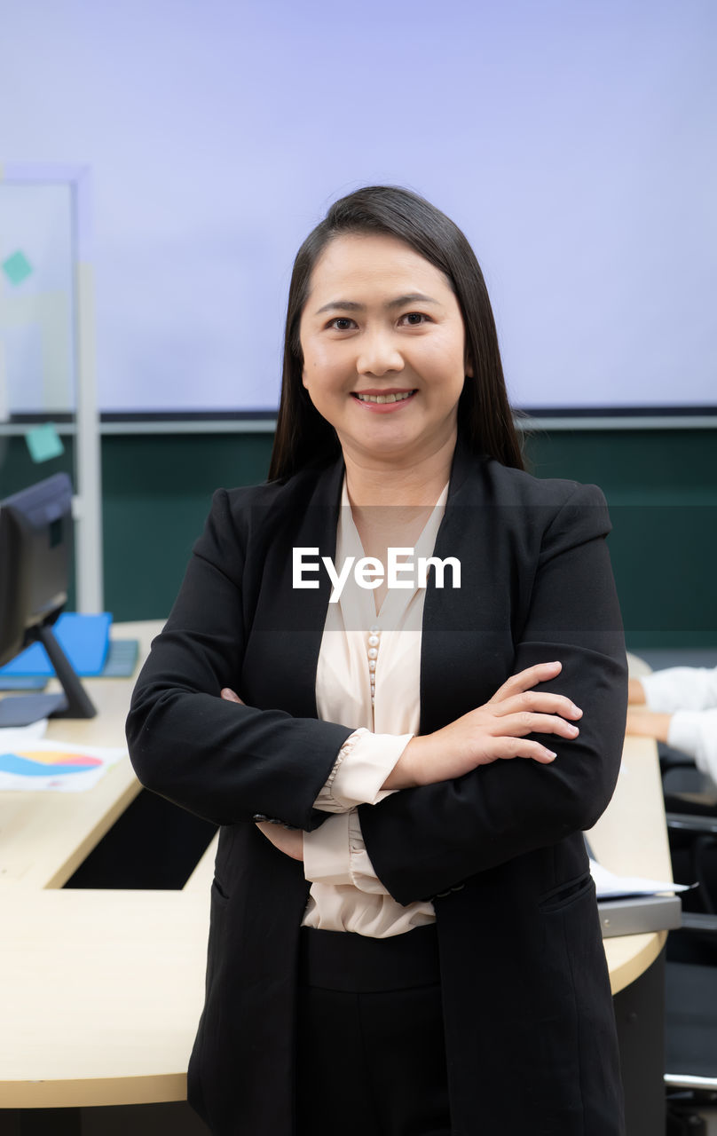 A woman in a black suit and white shirt is smiling and standing with her arms crossed.