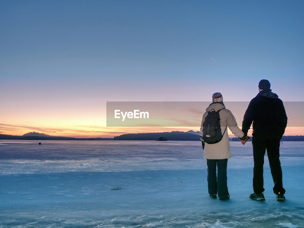 Couple has fun during winter walk on ice of frozen lake. evening hike at beach of pond. warm clothes