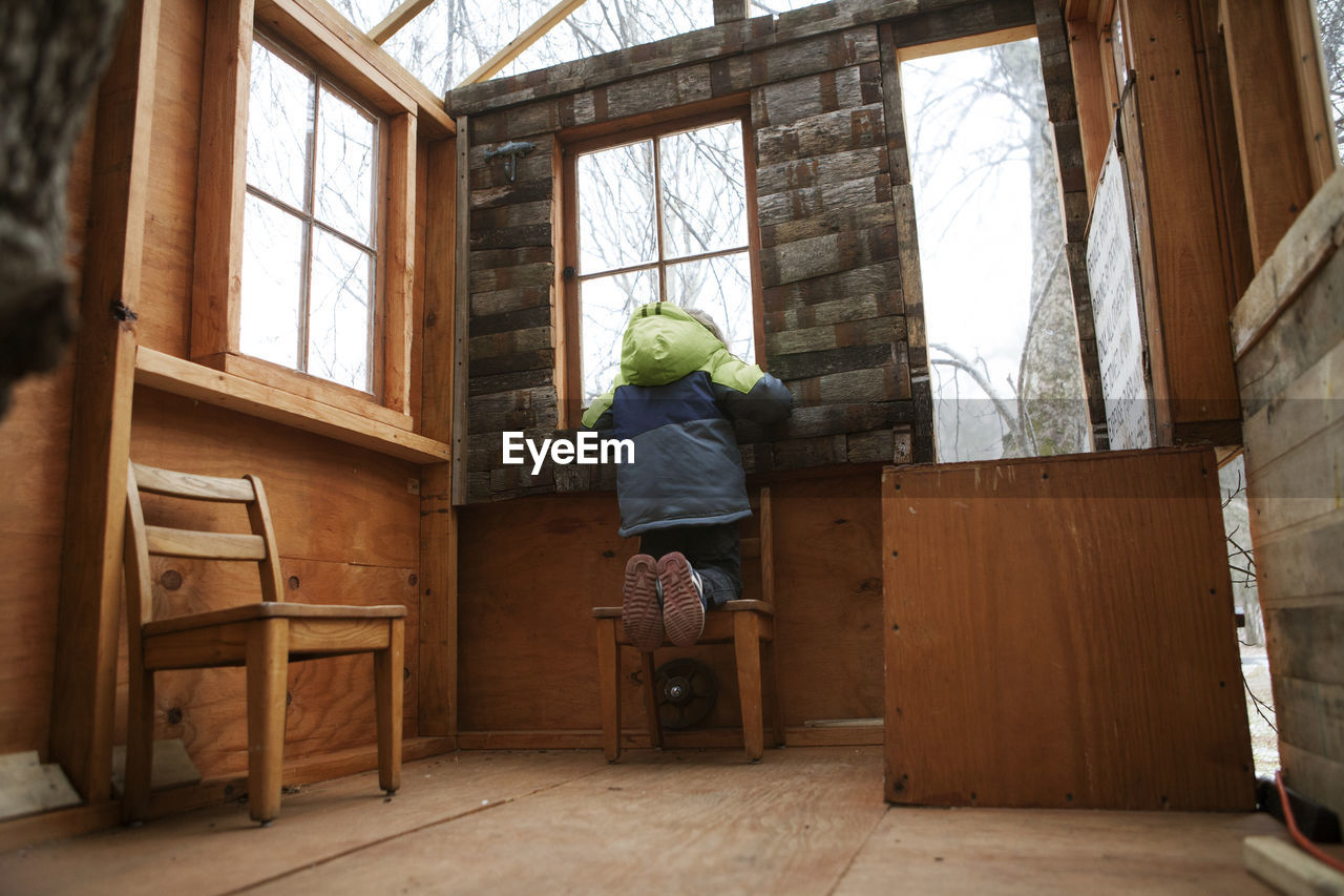 Rear view of boy looking through window while kneeling on chair in tree house