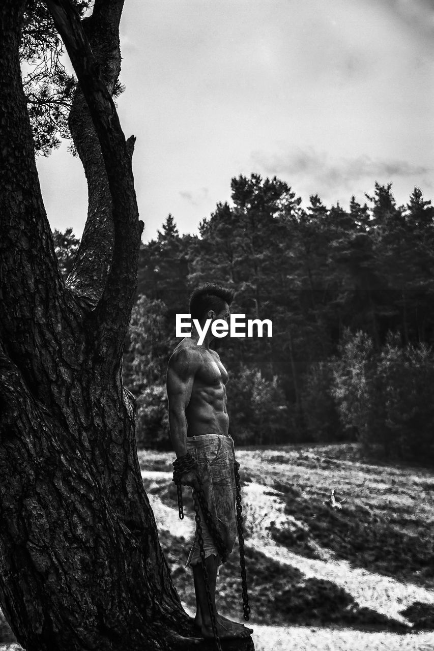 Side view of shirtless man standing by tree against sky