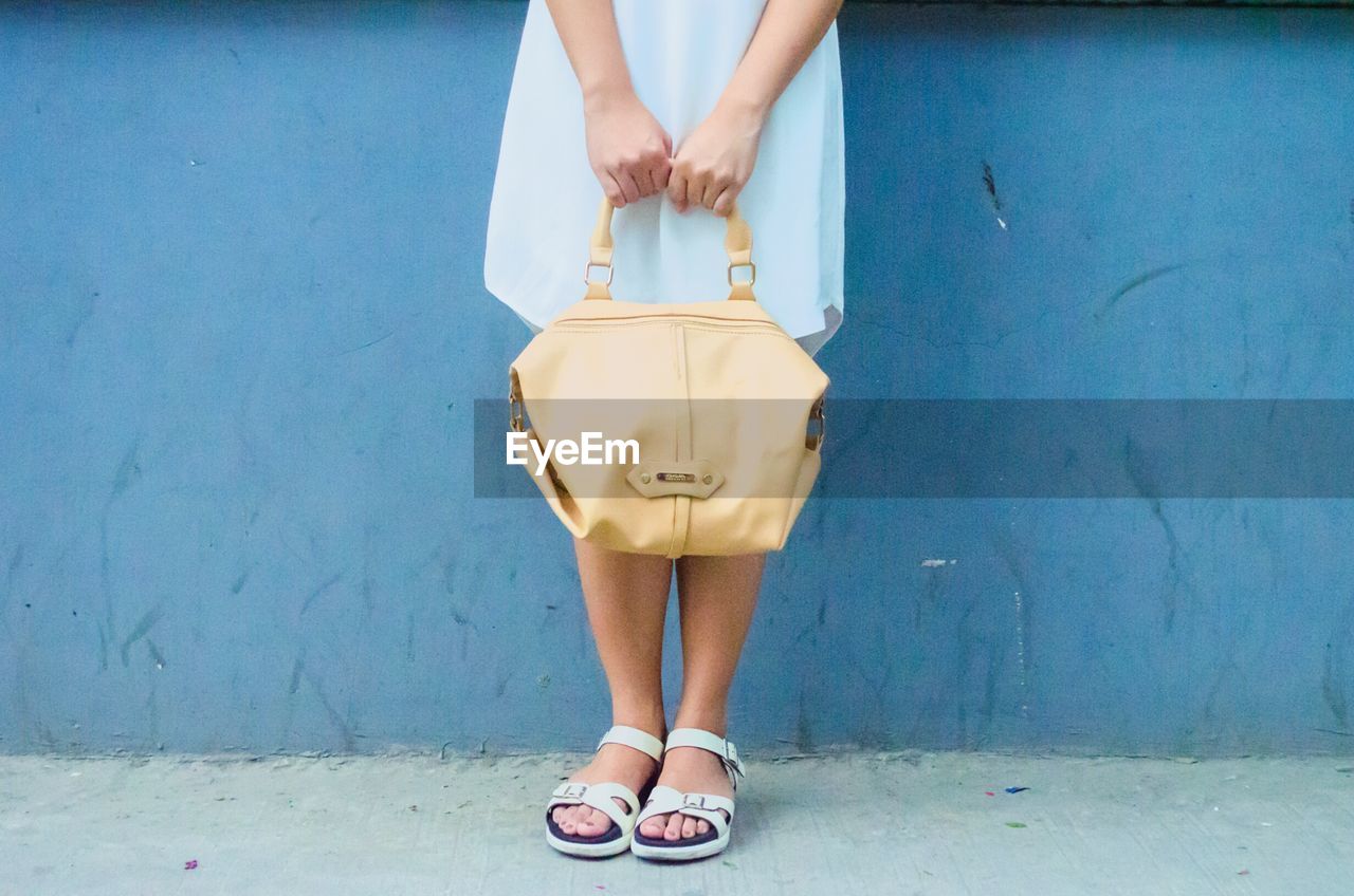 Low section of woman holding handbag while standing in front of blue wall