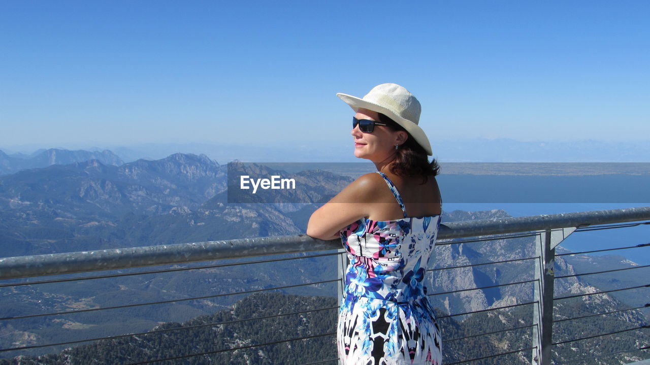 WOMAN STANDING ON RAILING AGAINST MOUNTAIN
