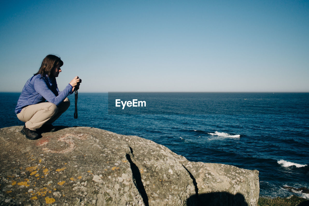 Side view of woman photographing while crouching on rocky shore