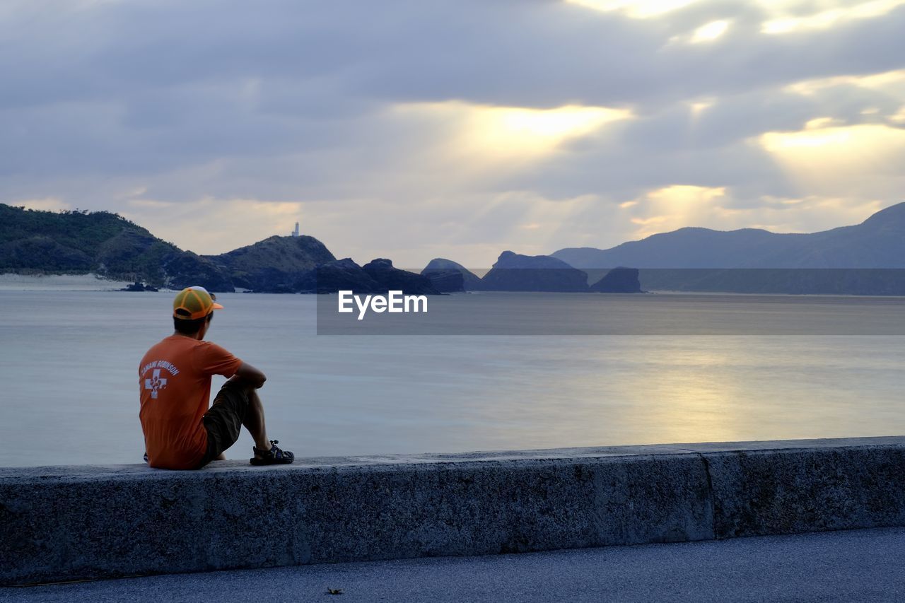 MAN SITTING ON LAKE AGAINST MOUNTAINS