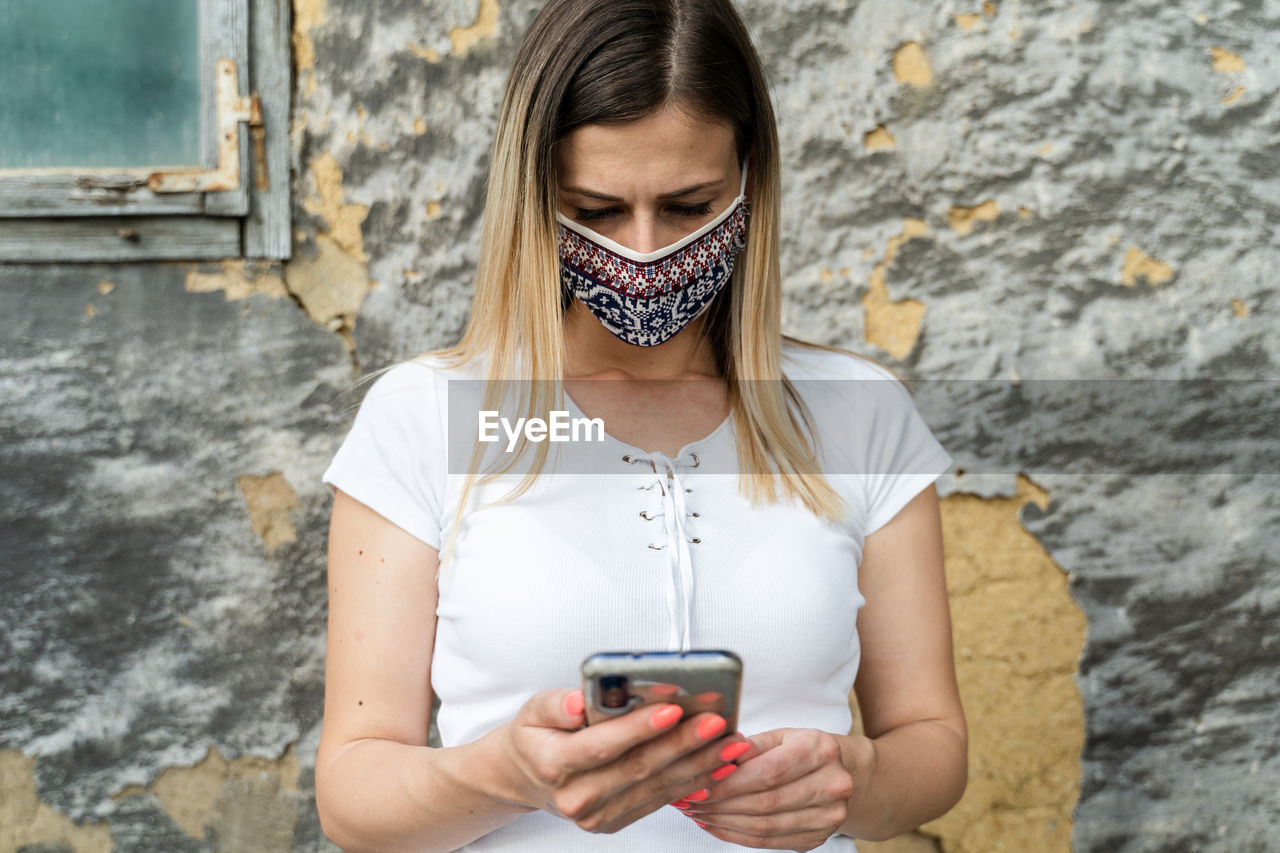 YOUNG WOMAN USING MOBILE PHONE AGAINST WALL