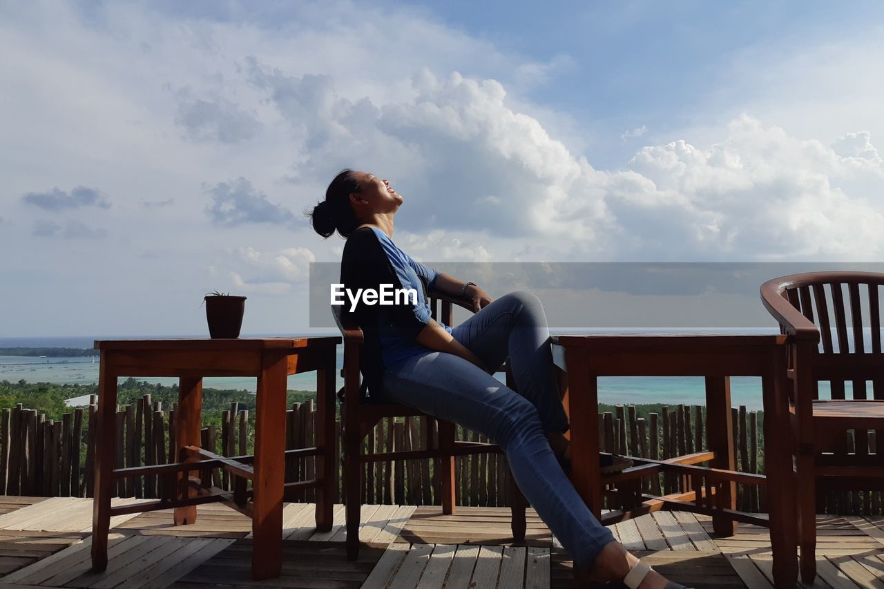 Side view of smiling woman sitting on chair against sky