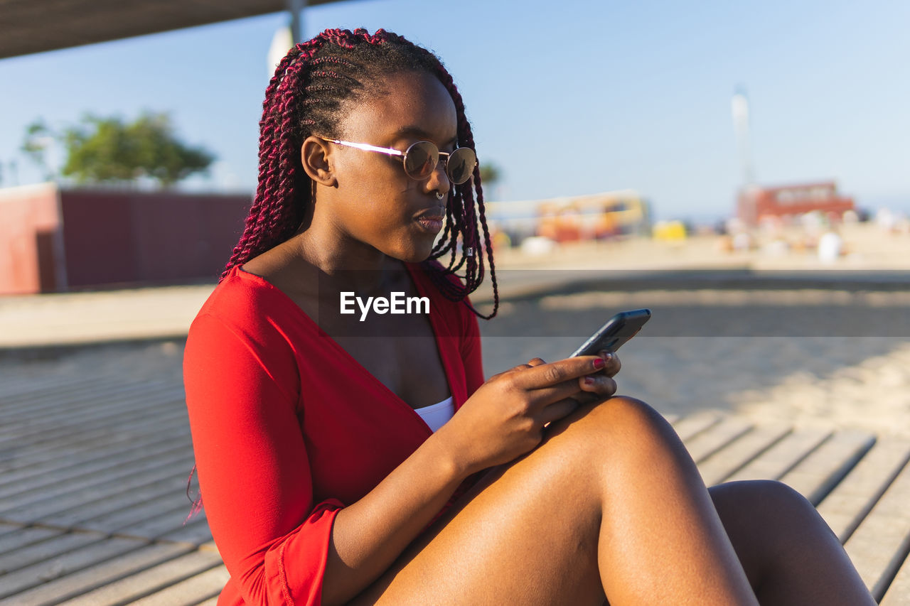 Focused african american young woman in trendy red dress and braids using phone while sitting at the beach