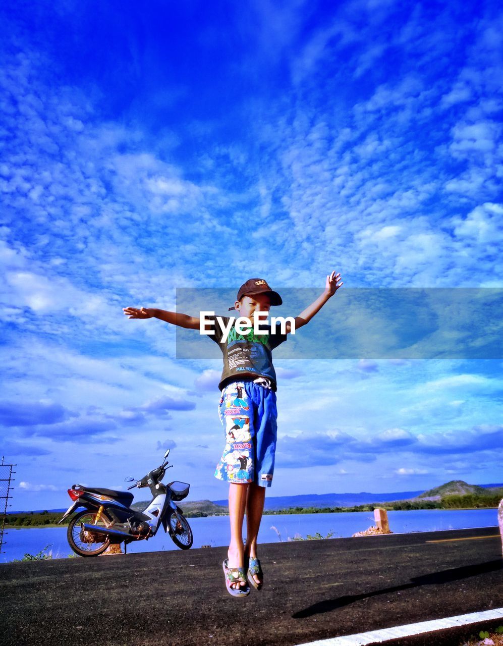 Full length of boy with arms outstretched jumping on road against sky