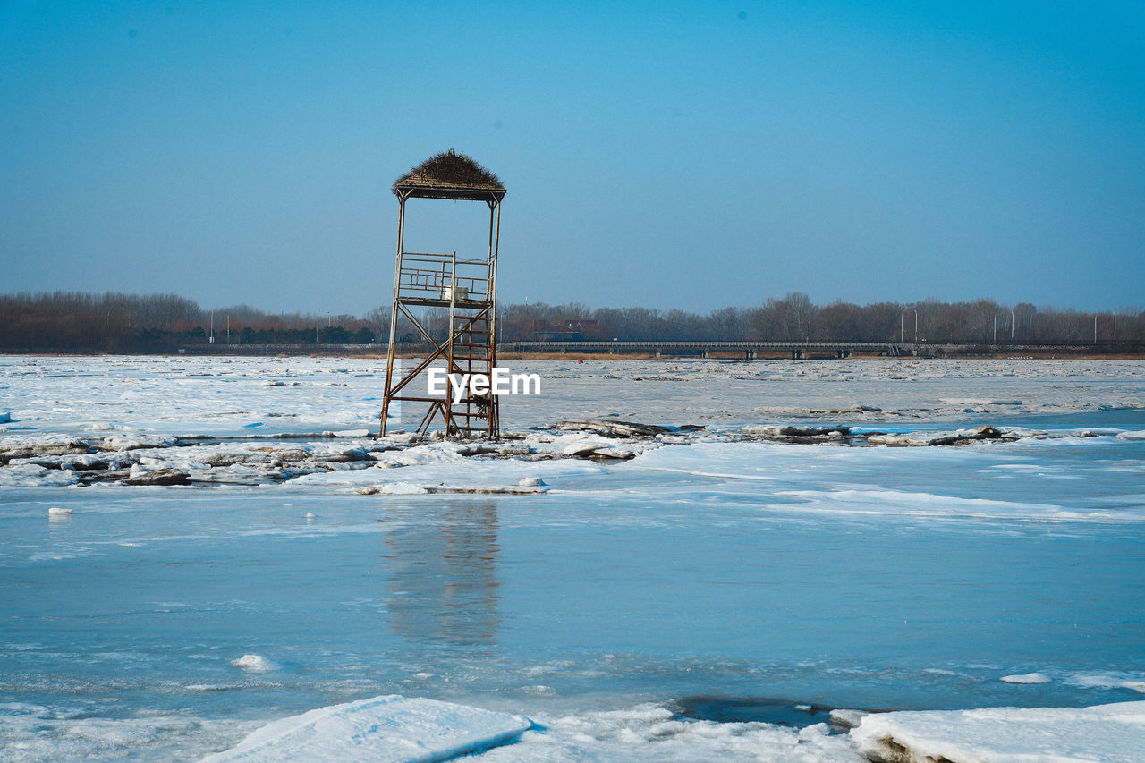 winter, water, ice, snow, cold temperature, sea, sky, nature, shore, reflection, ocean, no people, frozen, blue, scenics - nature, architecture, land, environment, day, built structure, tranquility, clear sky, tower, beauty in nature, tranquil scene, outdoors, freezing, wave, landscape, hut, beach, non-urban scene, coast