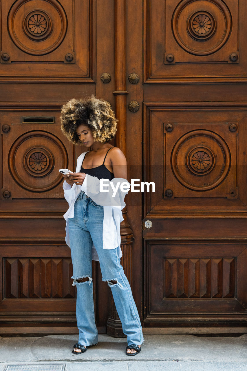 Full body of african american female in stylish casual outfit standing near entrance and surfing cellphone