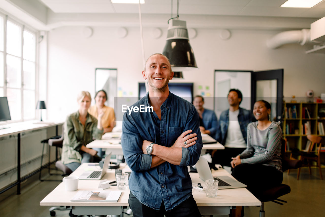 Portrait of smiling male entrepreneur standing with arms crossed and employees sitting at office desk