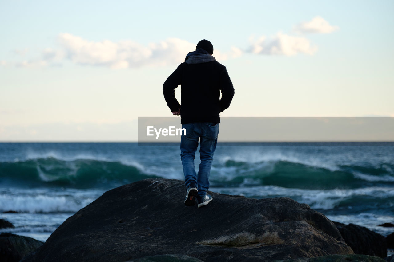 REAR VIEW OF MAN STANDING ON ROCK BY SEA